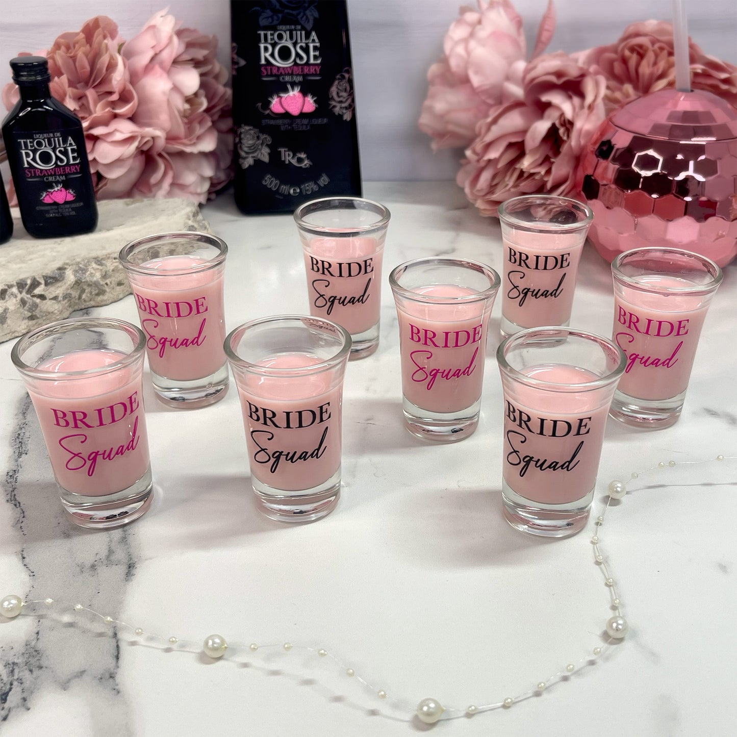 Pink shot glasses with 'Bride Squad' text on a marble surface with flowers and Tequila Rose bottle.
