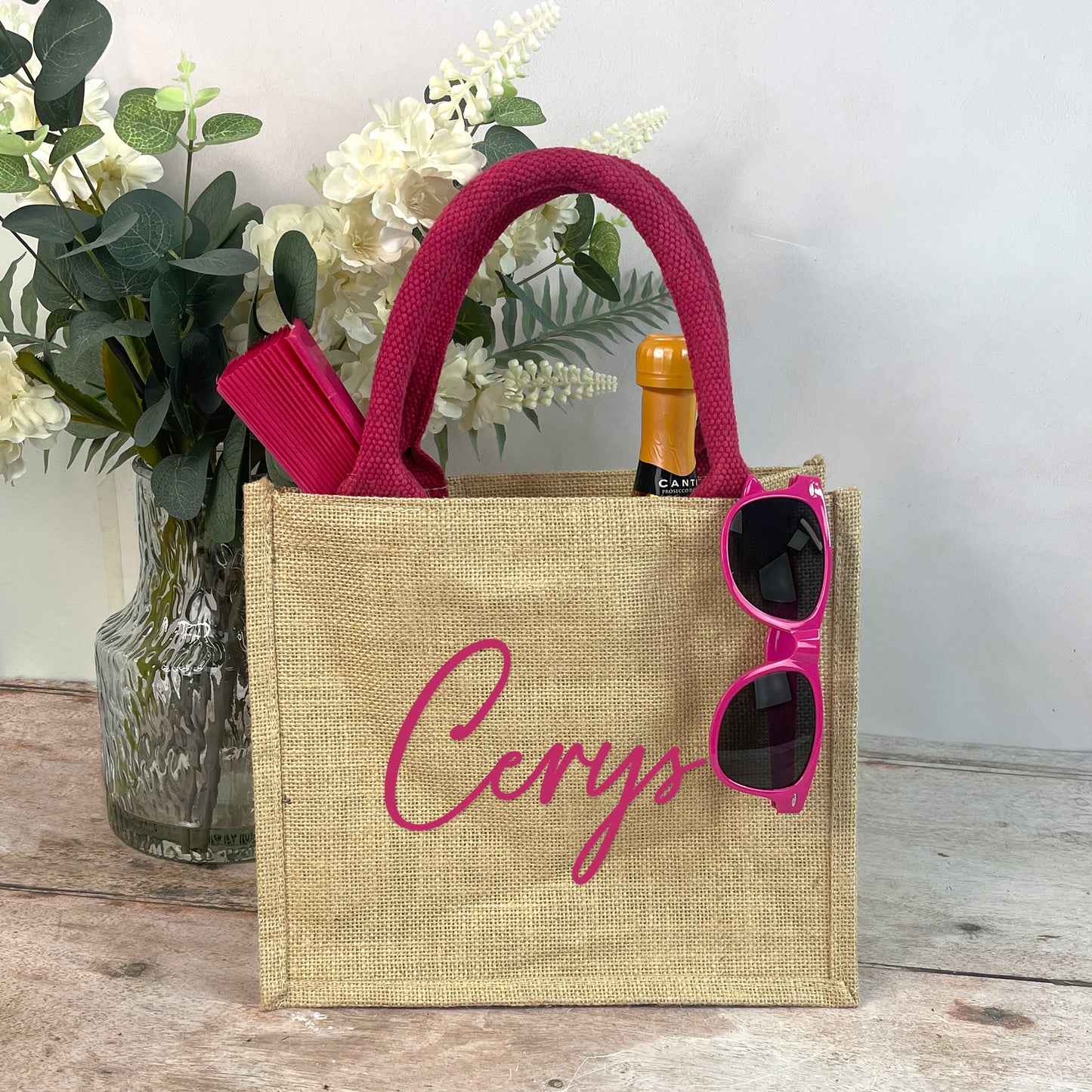 Jute bag with 'Cerys' branding, sunglasses, and a bottle on a wooden surface with flowers in the background.