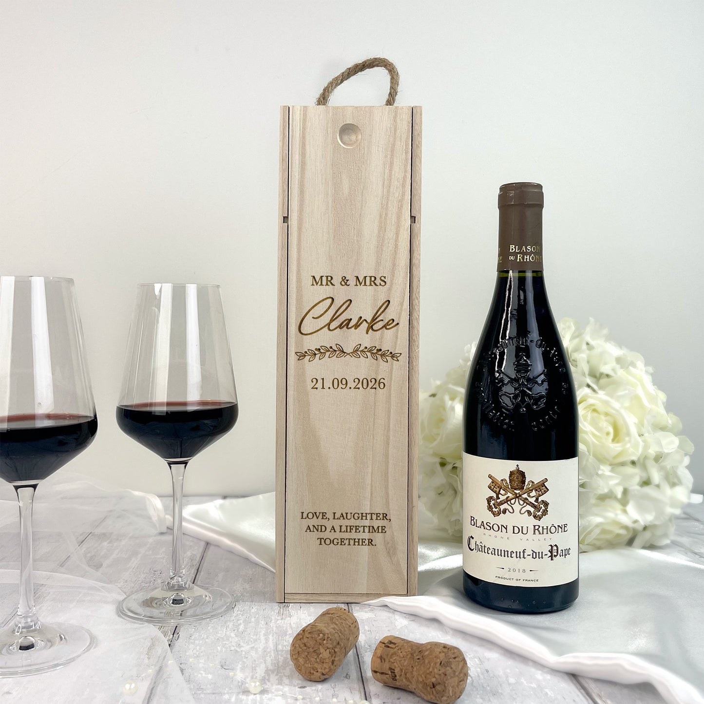 Wine bottle with wooden gift box, glasses, and flowers on a light surface.