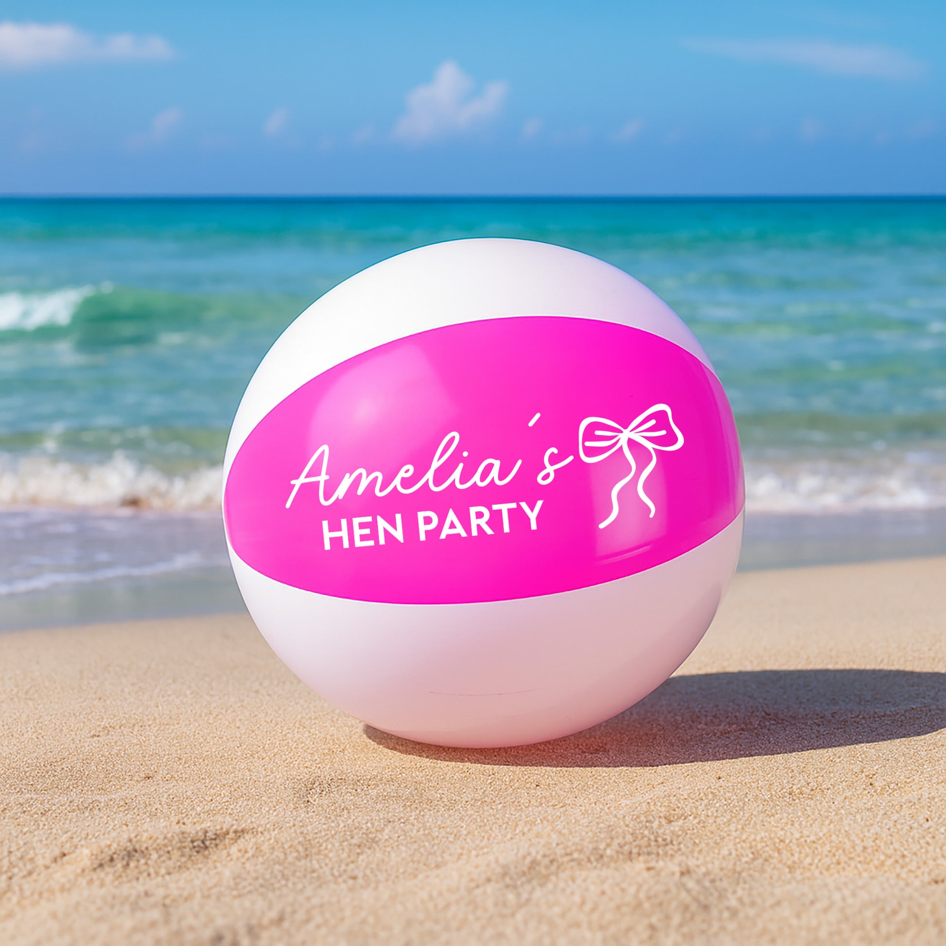A pink and white beach ball printed with 'Amelia's HEN PARTY' and a bow graphic, sitting on a sandy beach with the ocean and blue sky in the background.