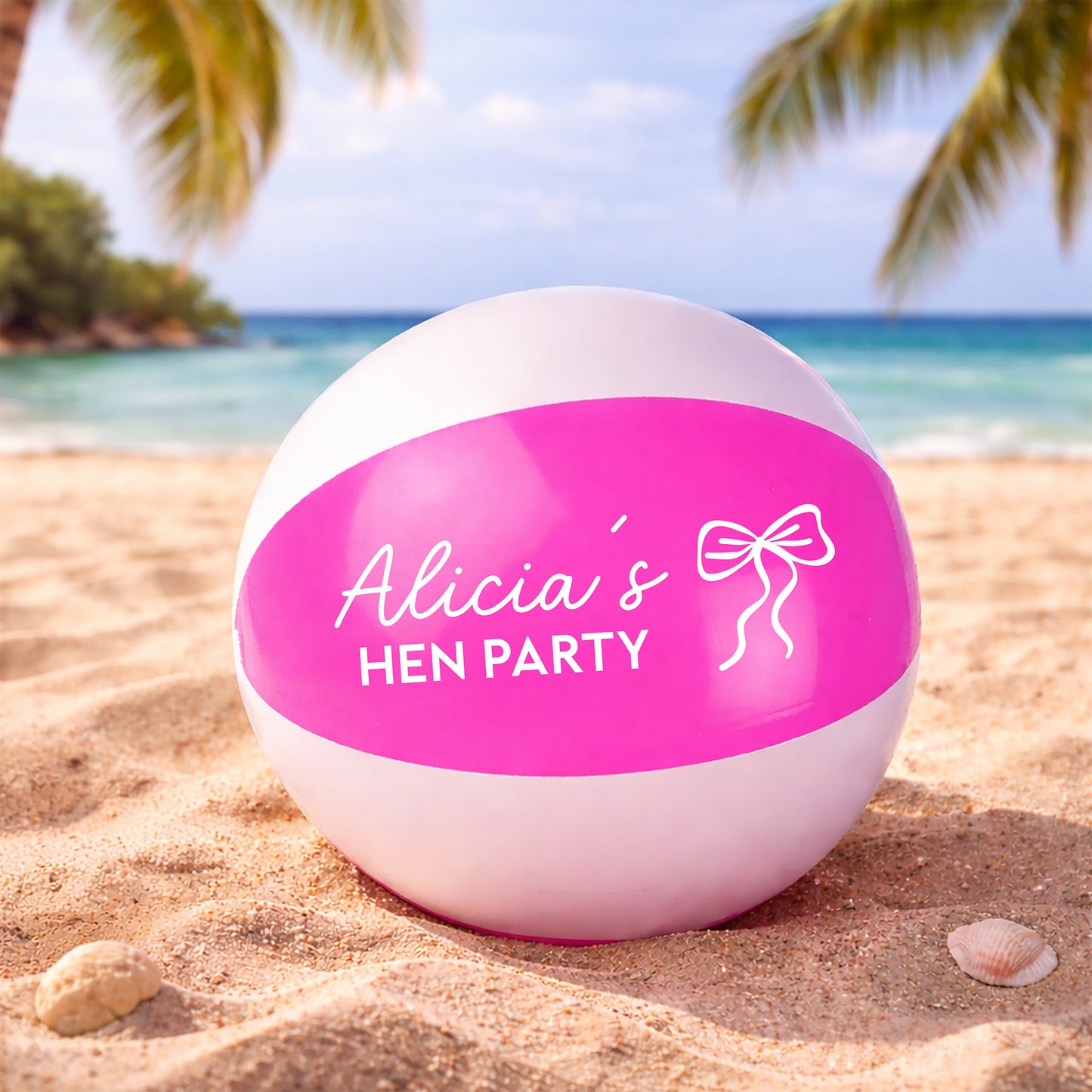 A pink and white beach ball printed with 'Alicia's HEN PARTY' and a bow graphic, resting on a sandy beach with a tropical ocean and palm trees in the background.
