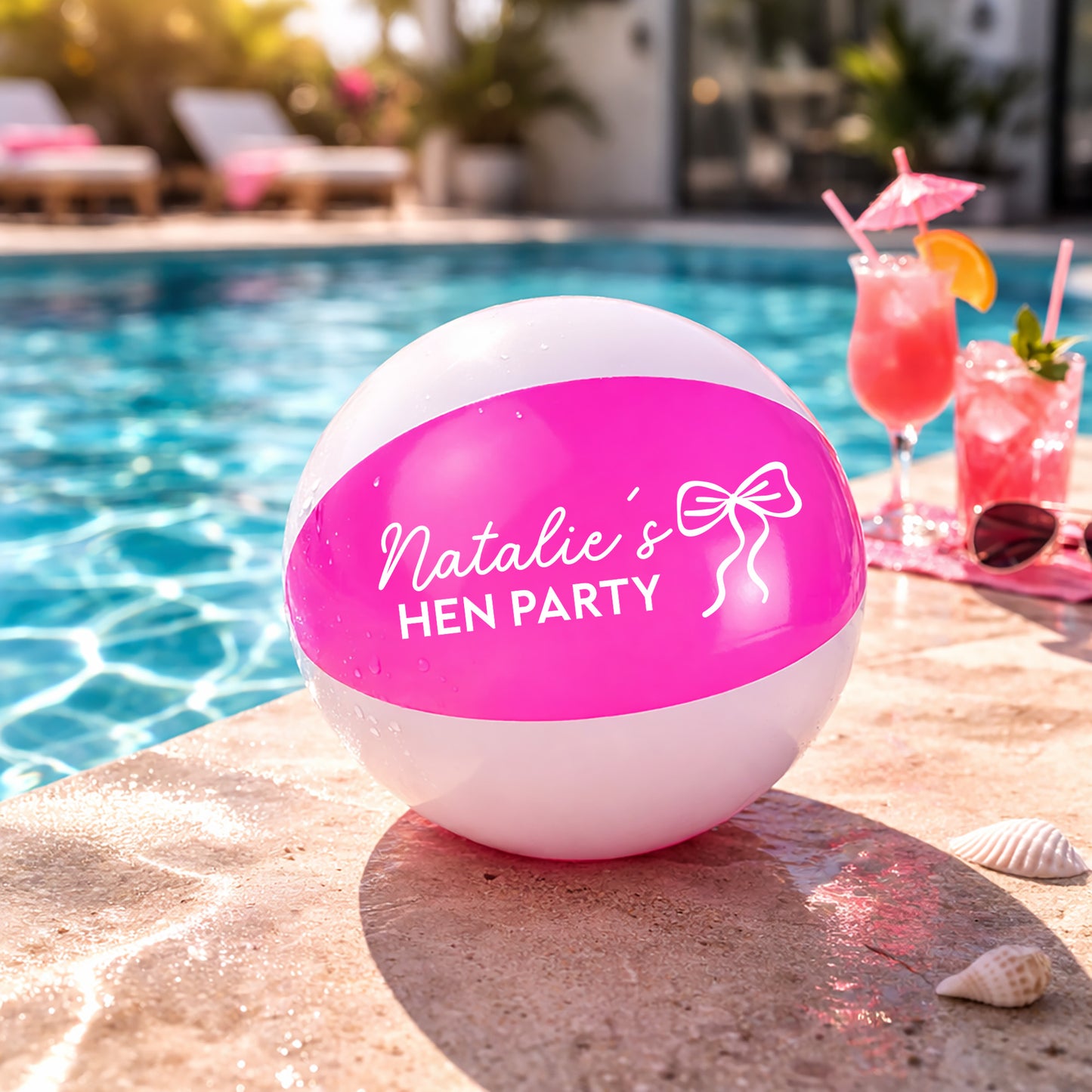 A white and bright pink beach ball printed with 'Natalie's HEN PARTY' sits on a stone poolside surface, with a swimming pool, cocktails, and seashells in the background.