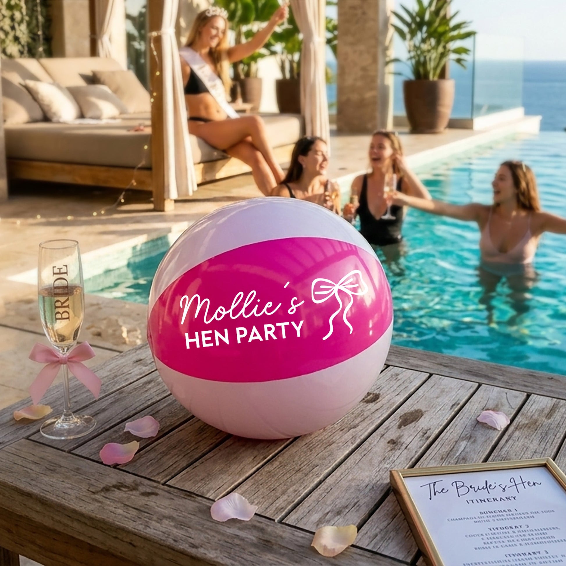 A pink and white beach ball printed with 'Mollie's HEN PARTY' sits on a wooden table next to a champagne flute and a party itinerary card, with a pool party in the background.