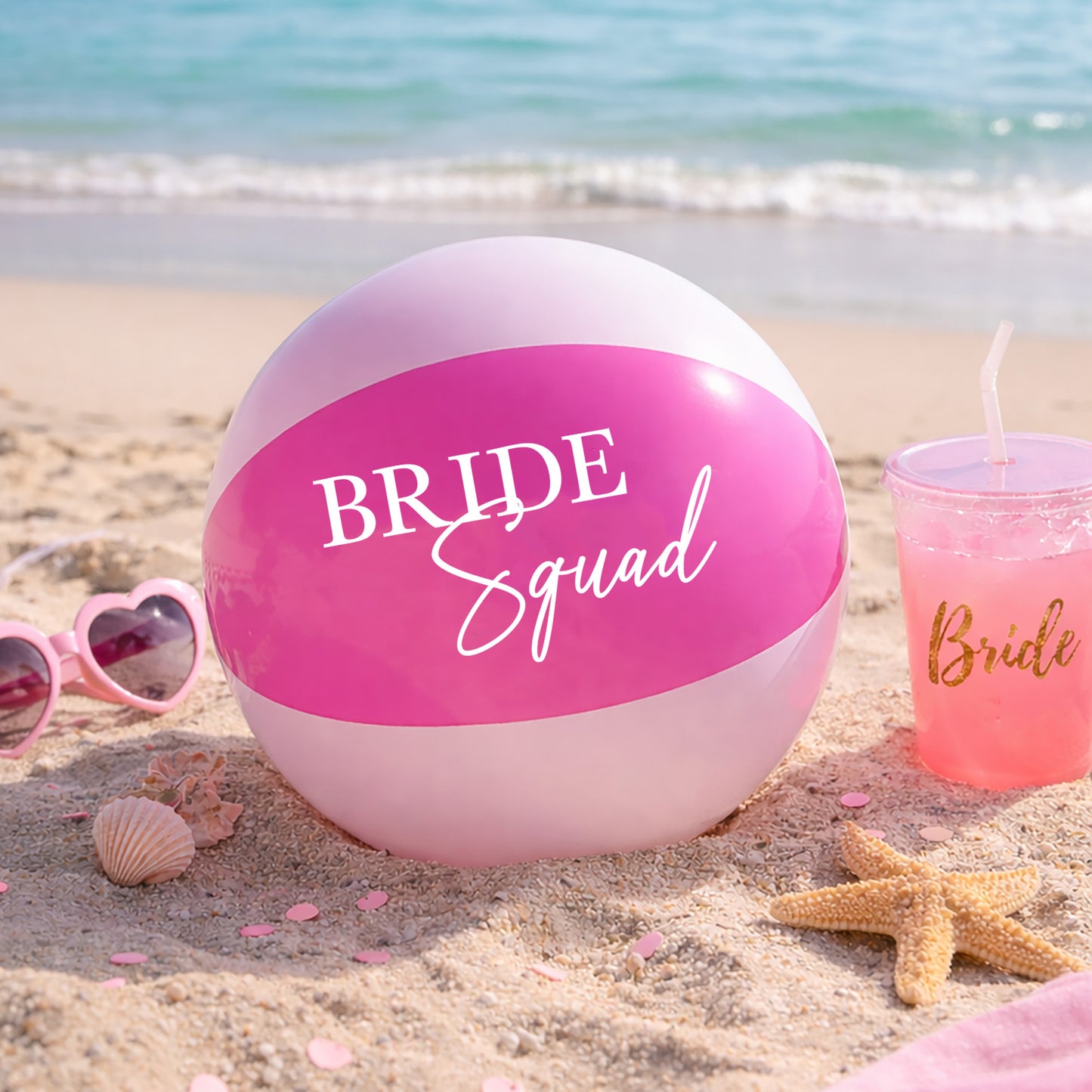 Pink beach ball with 'Bride Squad' text on a sandy beach with sunglasses and a drink.