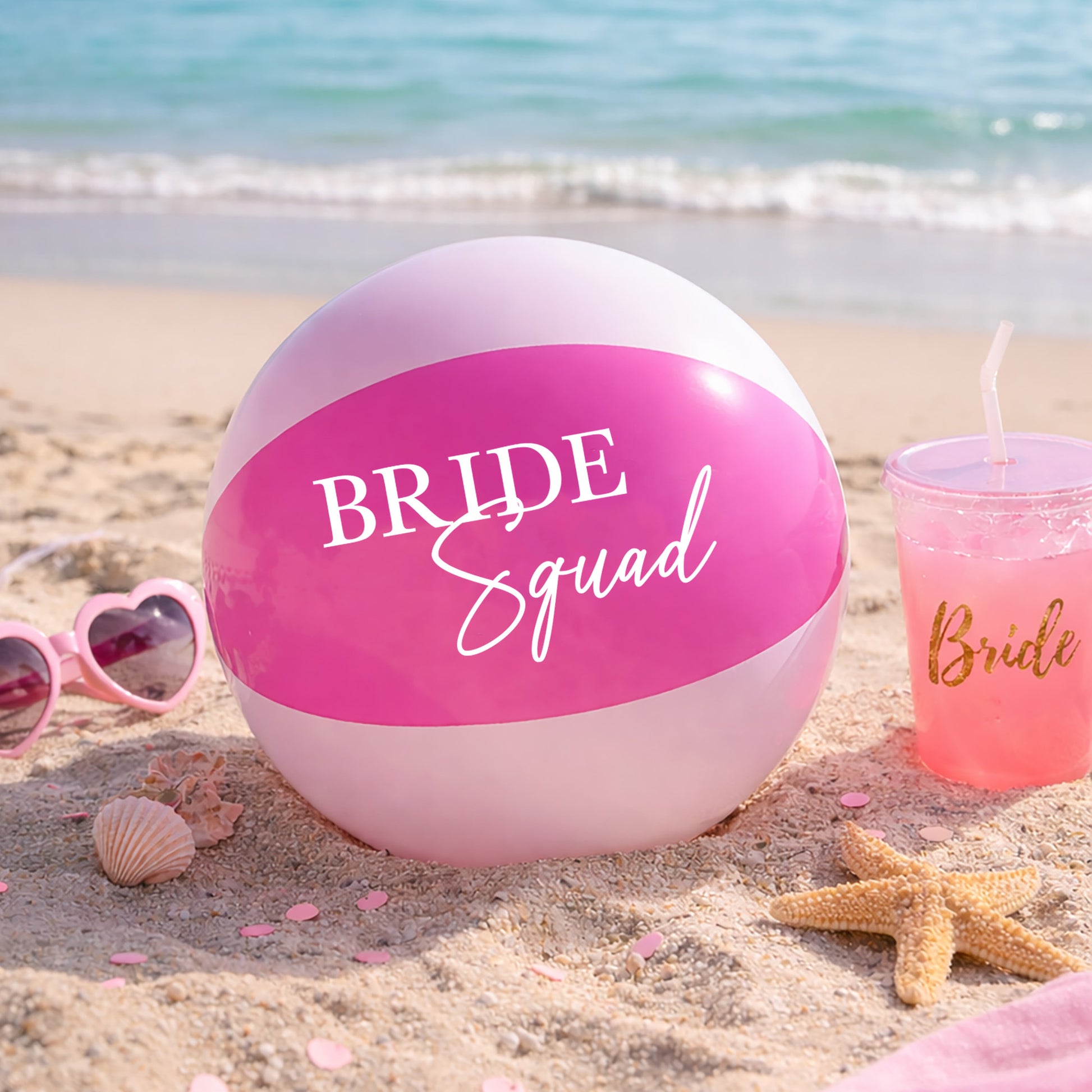 Pink beach ball with 'Bride Squad' text on a sandy beach with sunglasses and a drink.