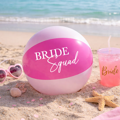 Pink beach ball with 'Bride Squad' text on a sandy beach with sunglasses and a drink.