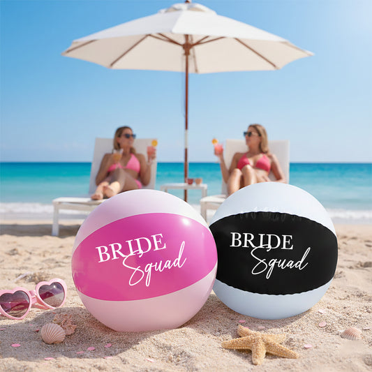 Two beach balls labeled 'Bride Squad' on a sandy beach with ocean and umbrellas in the background.