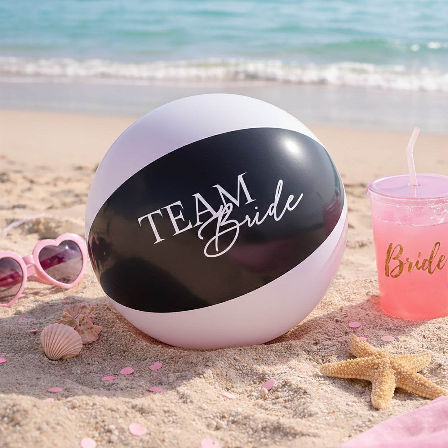 Beach ball with 'Team Bride' text on a sandy beach with sunglasses and a cup.