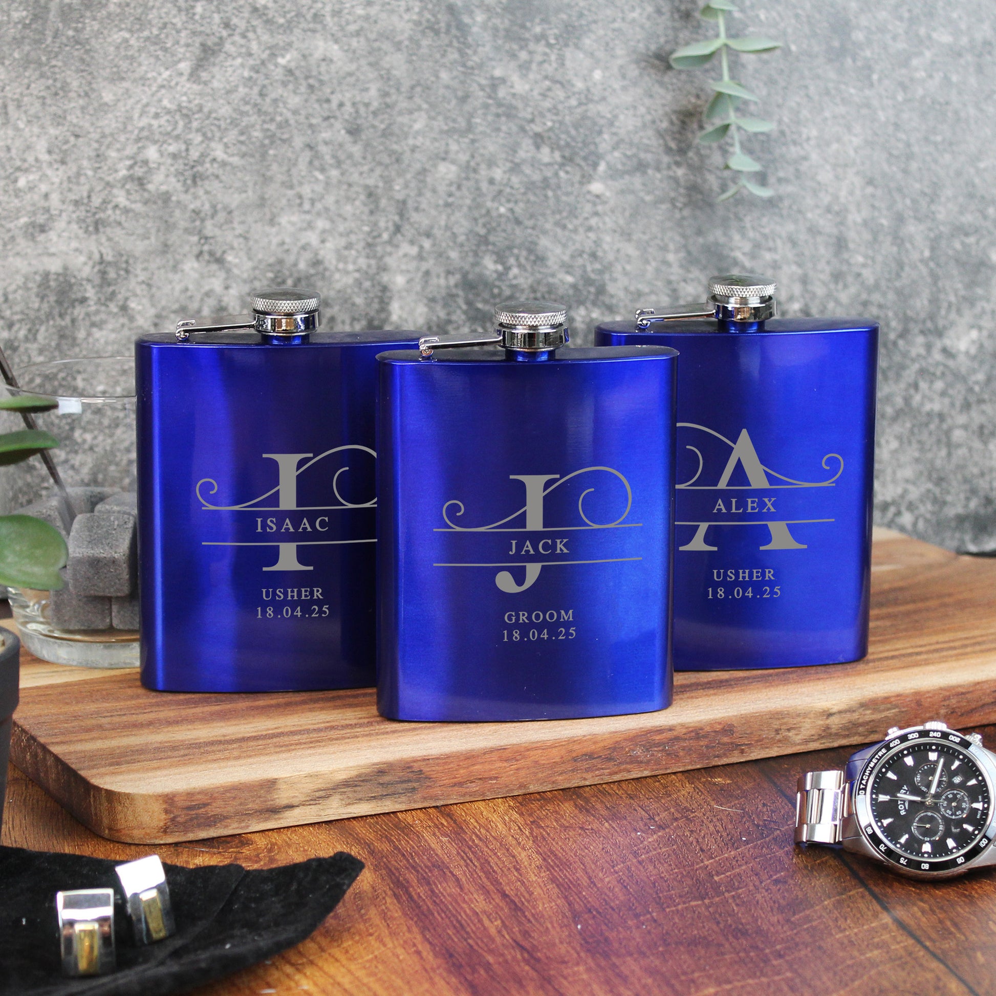 Three blue flasks with engraved names on a wooden surface with a watch and cufflinks.