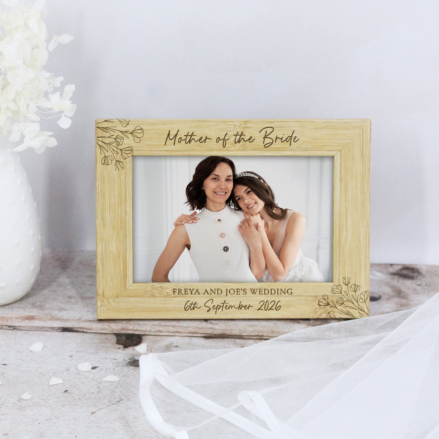 A personalized light wood picture frame with engraved floral corner details, displaying a wedding photo of a bride and groom. The top reads 'Mother of the Bride' and the bottom reads 'FREYA AND JOE'S WEDDING 6th September 2026'.