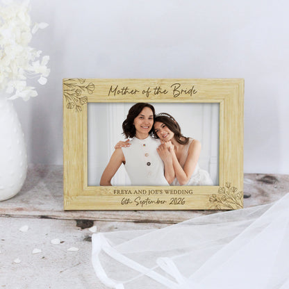 A personalized light wood picture frame with engraved floral corner details, displaying a wedding photo of a bride and groom. The top reads 'Mother of the Bride' and the bottom reads 'FREYA AND JOE'S WEDDING 6th September 2026'.