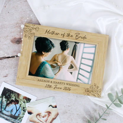 A personalized light wood photo frame engraved with 'Mother of the Bride' and wedding details, styled on a light surface with white fabric and instant photos.