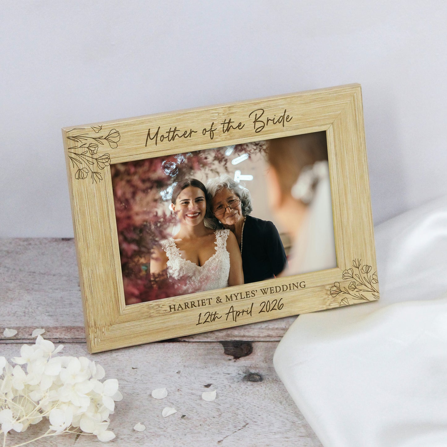 A rectangular wooden photo frame with engraved floral corner details and personalized wedding text reading 'Mother of the Bride' and 'Harriet & Myles Wedding 12th April 2026.' The frame is shown resting on a light surface with white fabric and small white flowers nearby.