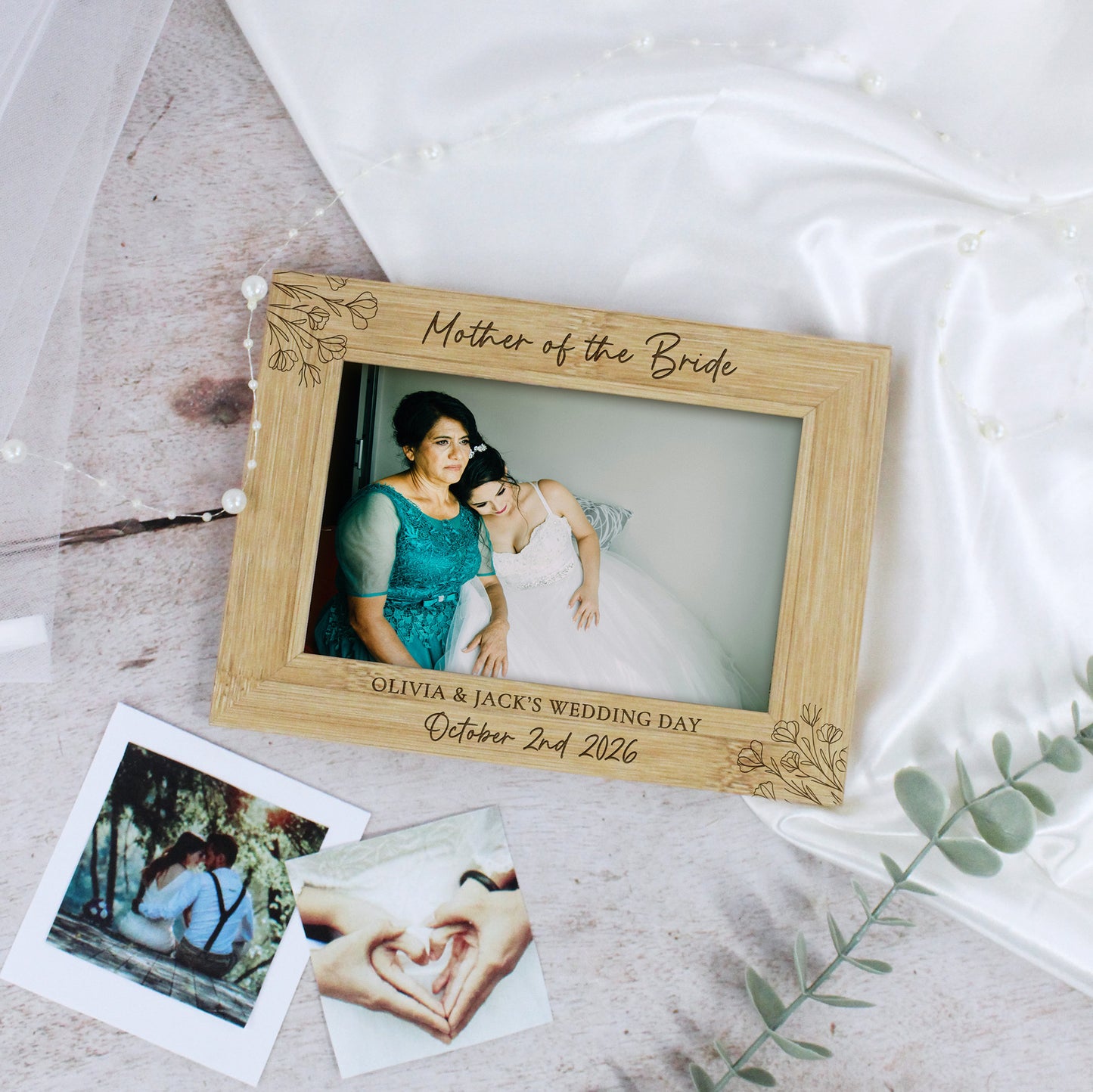 A personalized wooden picture frame engraved with 'Mother of the Bride' and wedding details, styled on a light surface with satin fabric, a tulle veil, and instant photos.