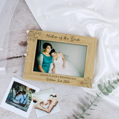 A personalized wooden picture frame engraved with 'Mother of the Bride' and wedding details, styled on a light surface with satin fabric, a tulle veil, and instant photos.