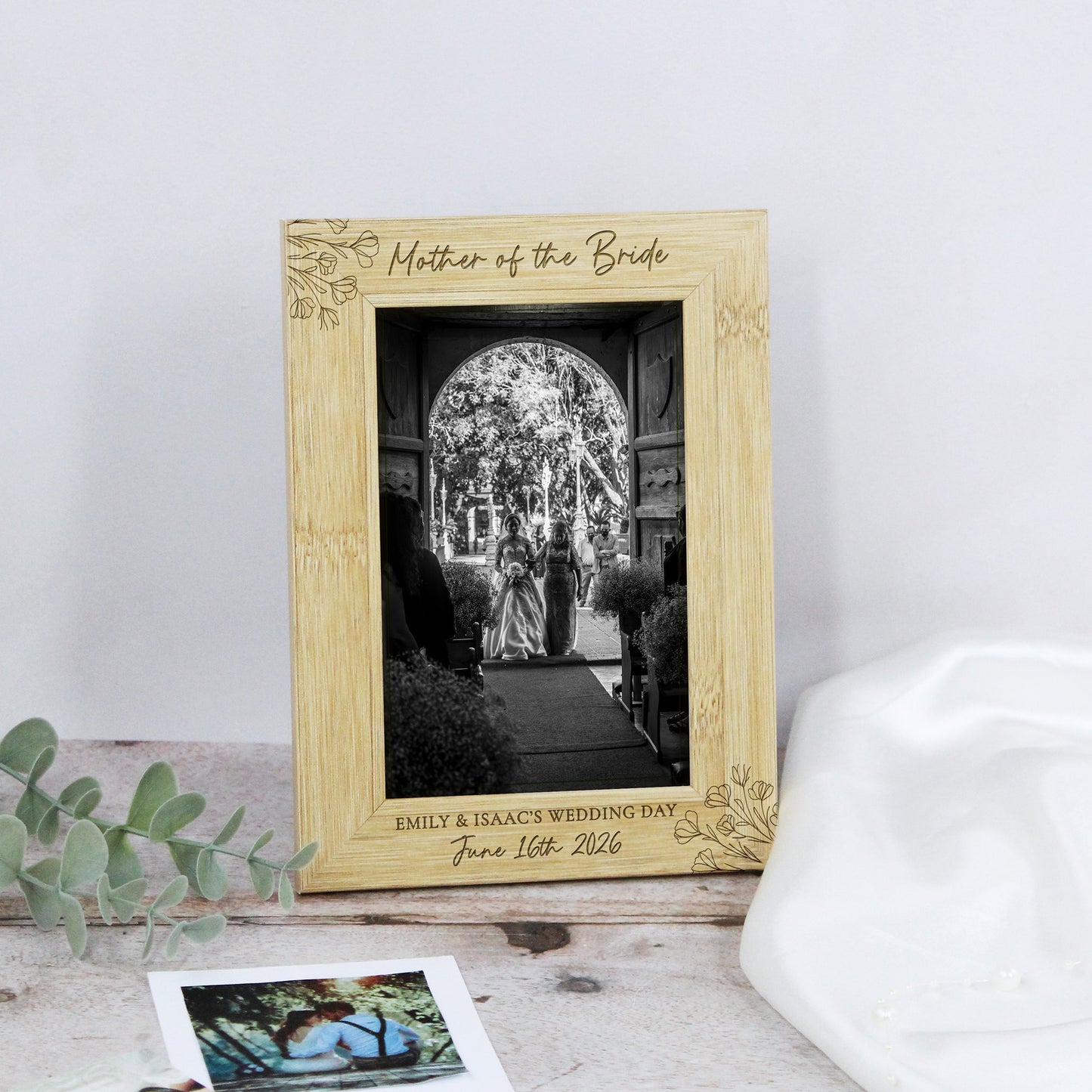 A personalized light wood picture frame engraved with 'Mother of the Bride' and wedding details, displayed on a rustic surface with eucalyptus leaves and a white hat nearby.