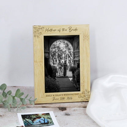 A personalized light wood picture frame engraved with 'Mother of the Bride' and wedding details, displayed on a rustic surface with eucalyptus leaves and a white hat nearby.