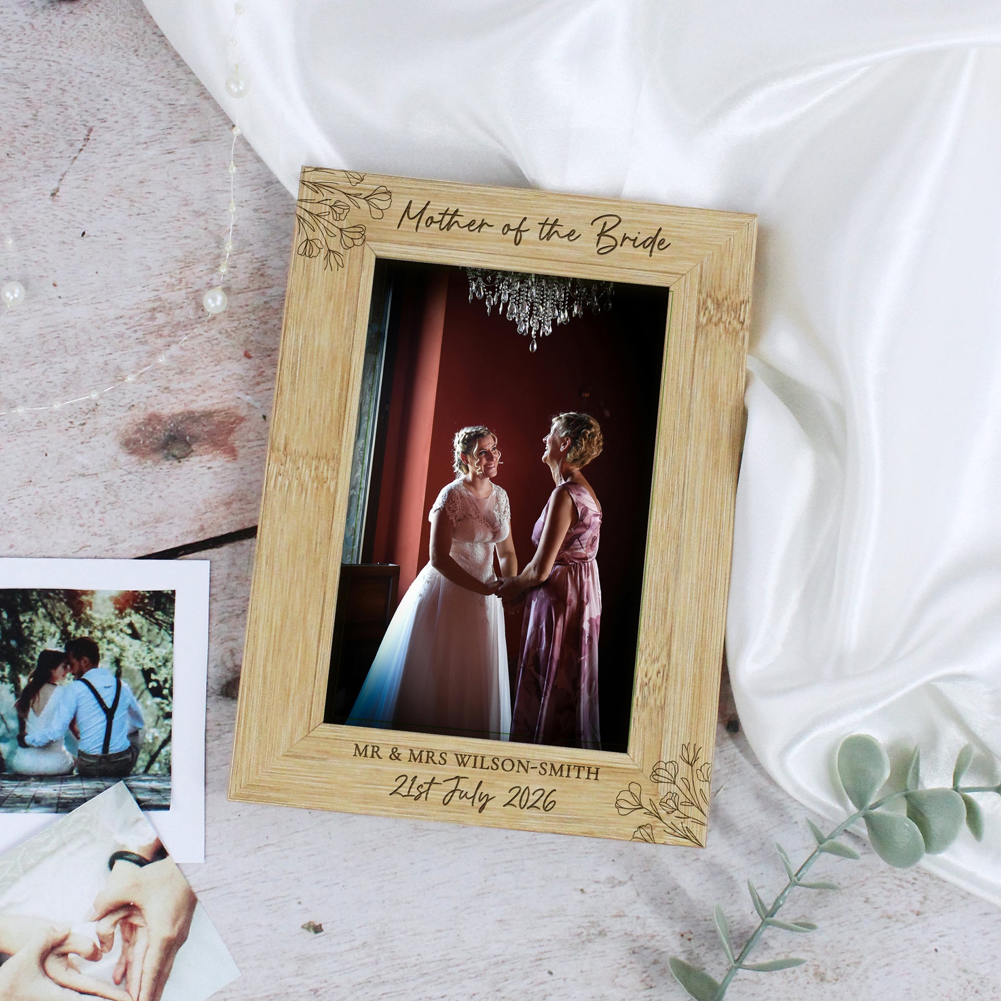 A personalized bamboo photo frame engraved with 'Mother of the Bride' and wedding details, styled on a rustic wooden surface with white satin fabric, a small instant photo, and eucalyptus leaves.