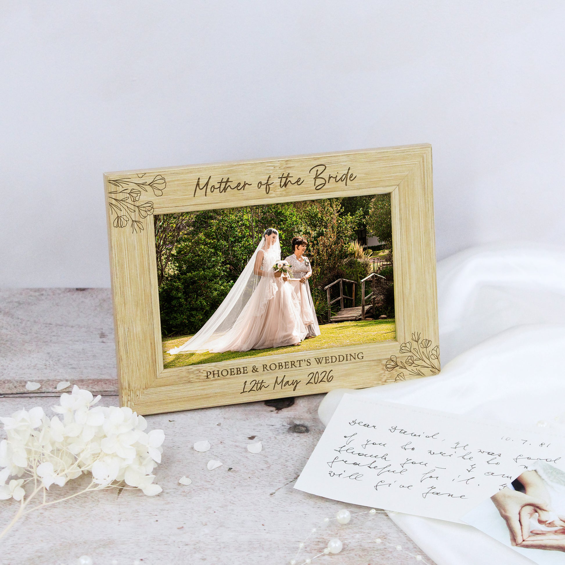 A personalized wooden wedding photo frame with engraved floral corner details and the text 'Mother of the Bride' and custom wedding details, styled on a rustic surface with dried white flowers and a handwritten card.