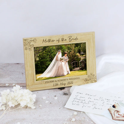 A personalized wooden wedding photo frame with engraved floral corner details and the text 'Mother of the Bride' and custom wedding details, styled on a rustic surface with dried white flowers and a handwritten card.