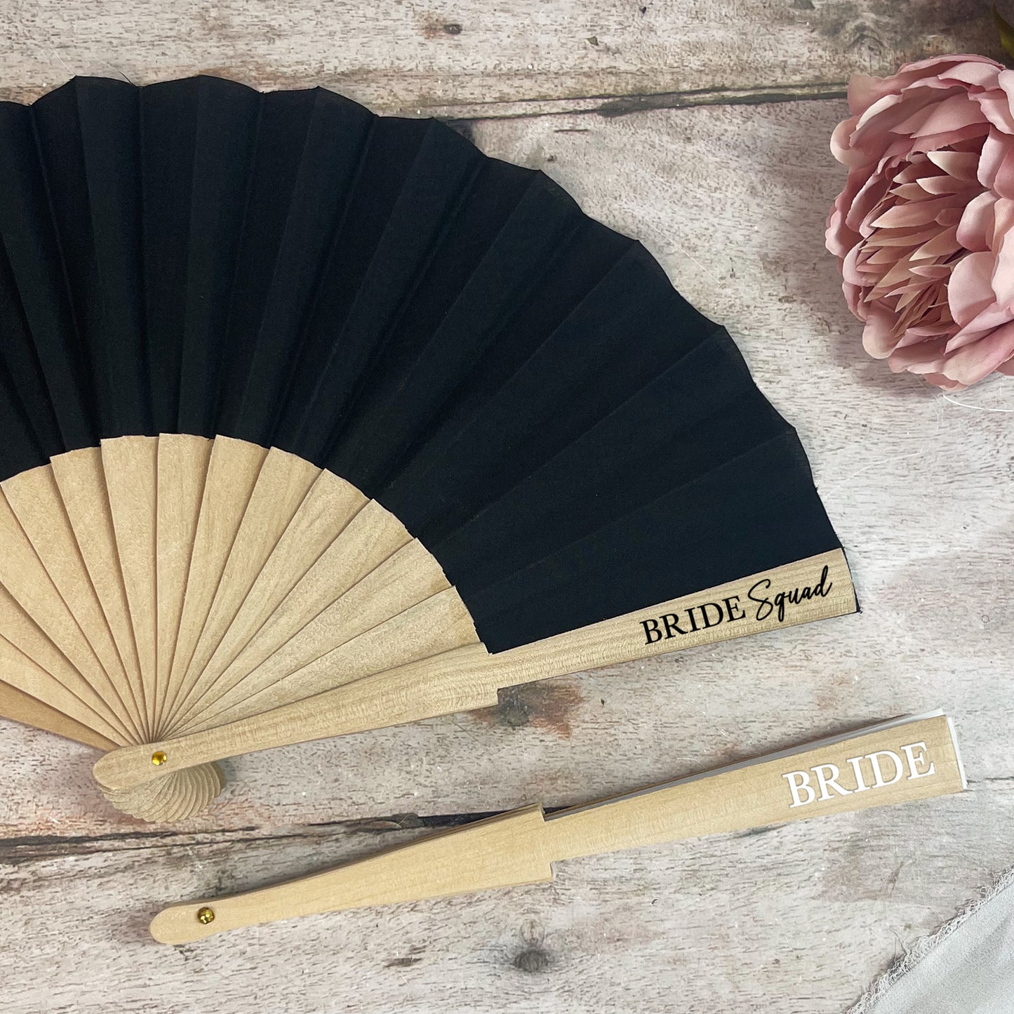 Black and gold fan with 'BRIDE Squad' and 'BRIDE' text on a wooden surface with pink flowers.