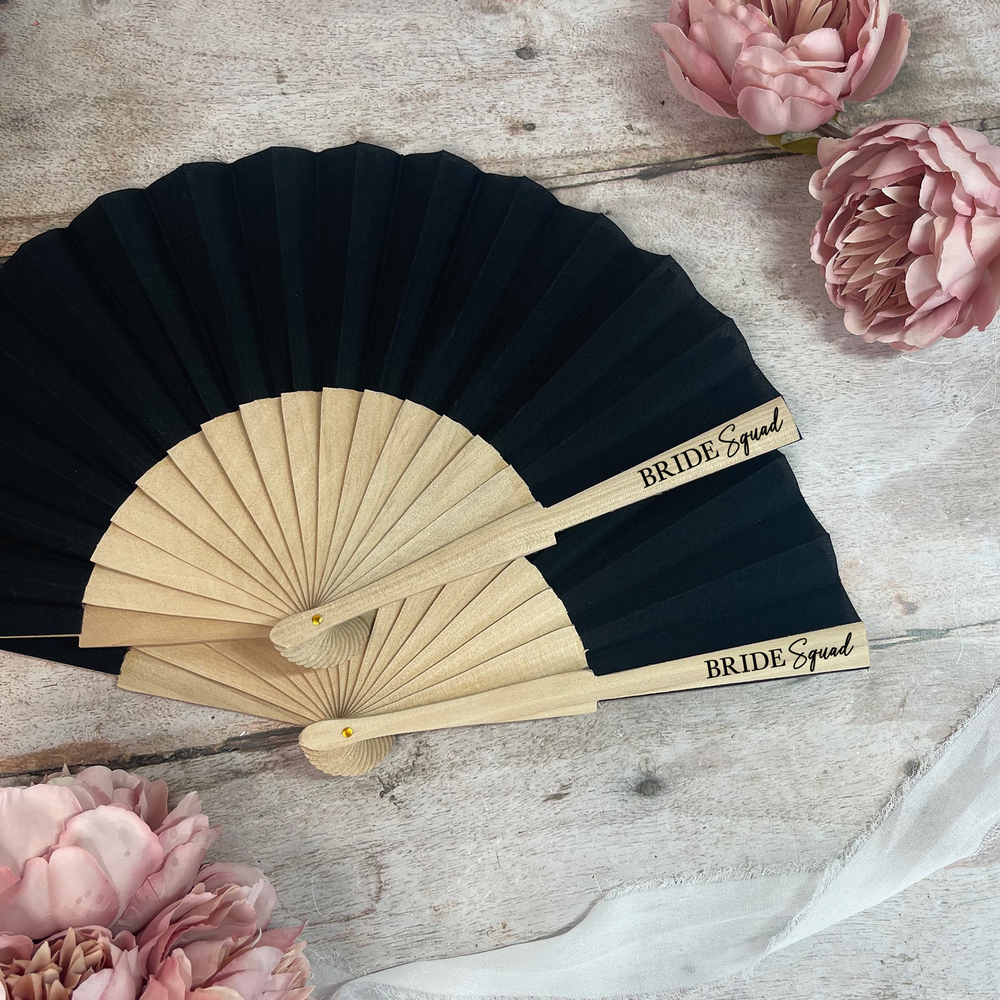 Black and beige hand fan with 'Bride Squad' text on a wooden surface with pink flowers.