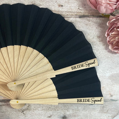 Black and beige hand fan with 'Bride Squad' text on a wooden surface with flowers.