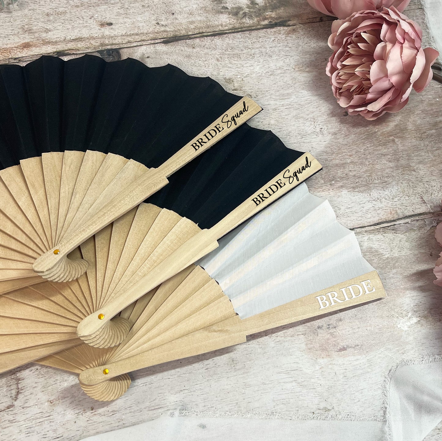Black and beige fans with 'Bride Squad' and 'Bride' text on a wooden surface with flowers.
