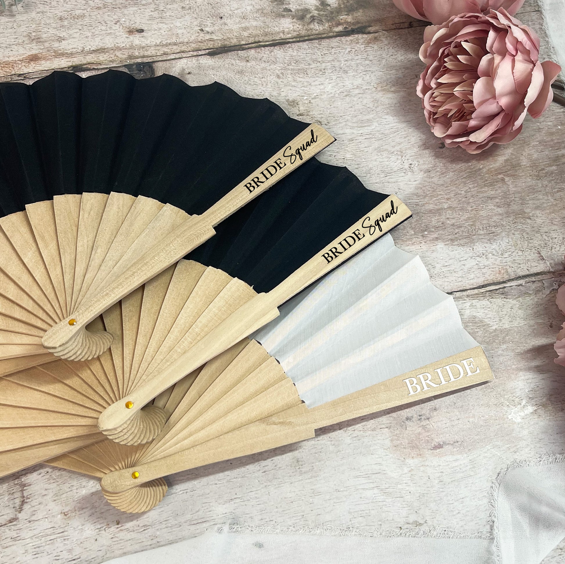 Black and beige fans with 'Bride Squad' and 'Bride' text on a wooden surface with flowers.