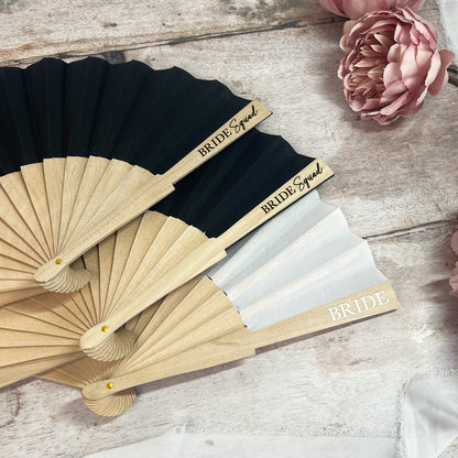 Black and beige fans with 'Bride Squad' and 'Bride' text on a wooden surface with flowers.