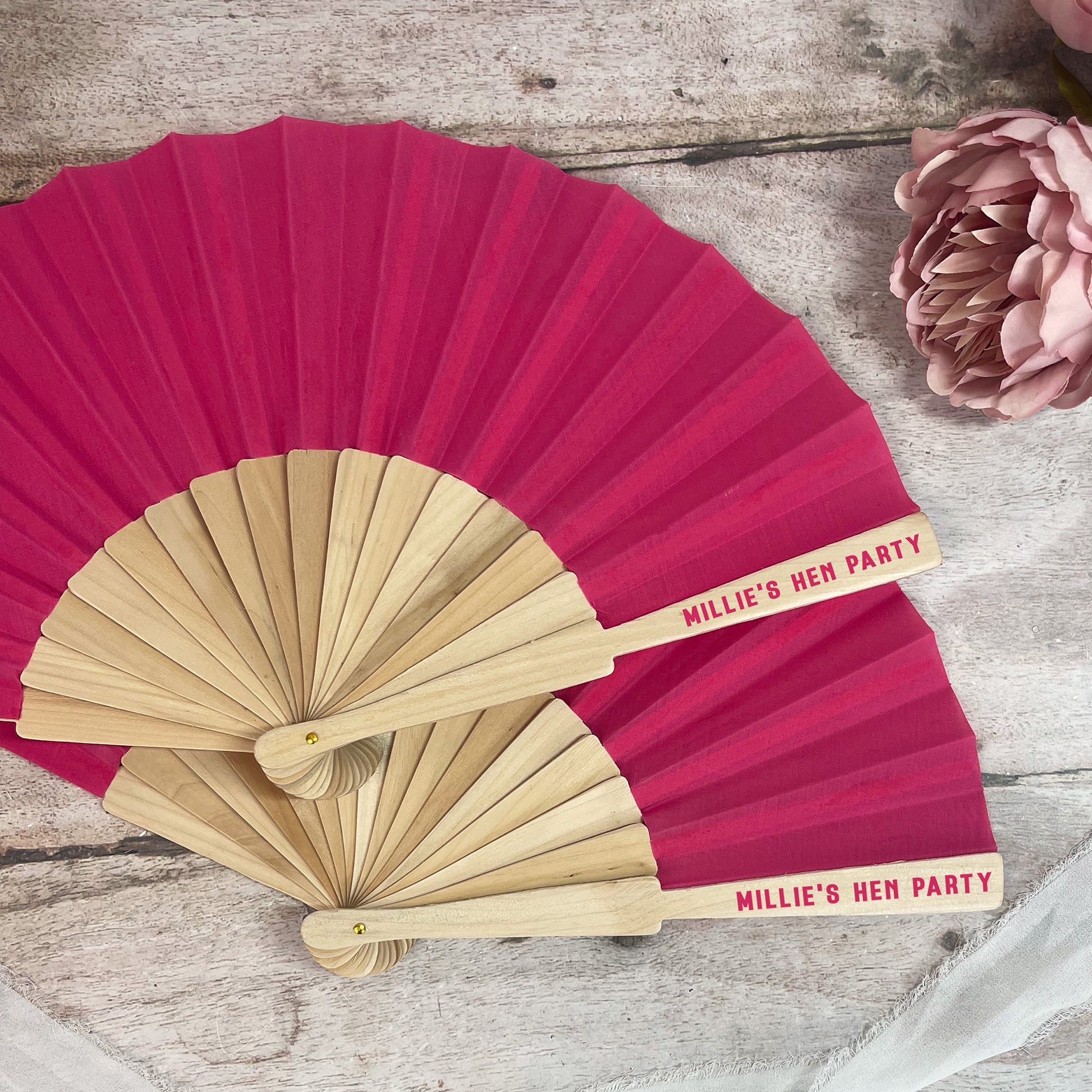 Two hand fans, one red and one beige, with 'Millie's Hen Party' text on a wooden surface.