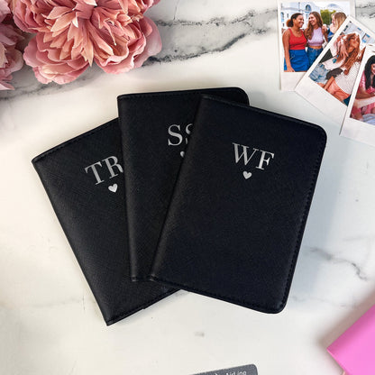 Three black passport holders with initials on a marble surface with flowers and photos.