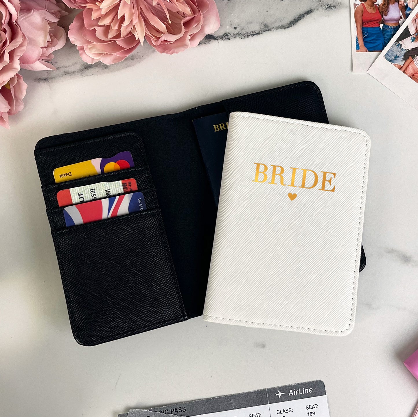 Black wallet with colorful cards, white passport holder with 'BRIDE' text, and airplane tickets on a marble surface with pink flowers.