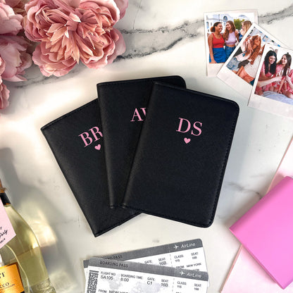 Three black passport holders with initials on a marble surface with flowers and travel items.