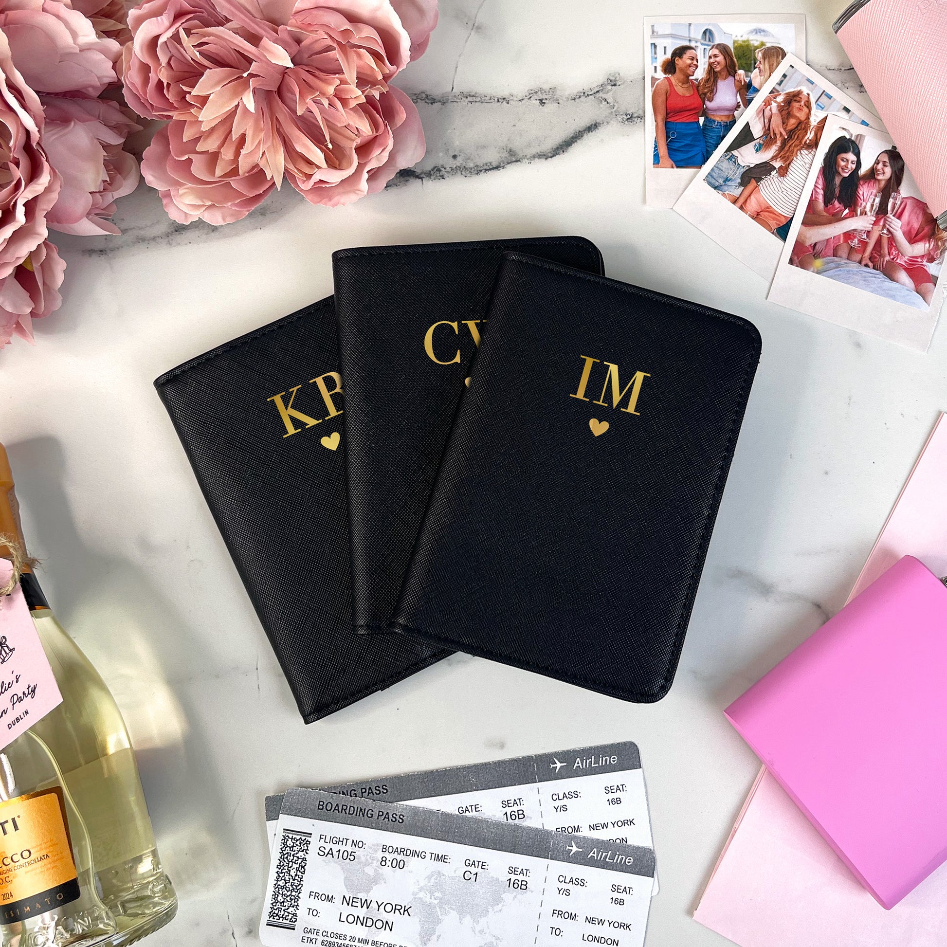 Three black passport holders with gold initials on a marble surface with flowers, wine, and photos.