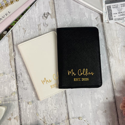 Two passport holders, one black and one beige, with 'Mr Collins EST. 2026' text on a wooden surface.