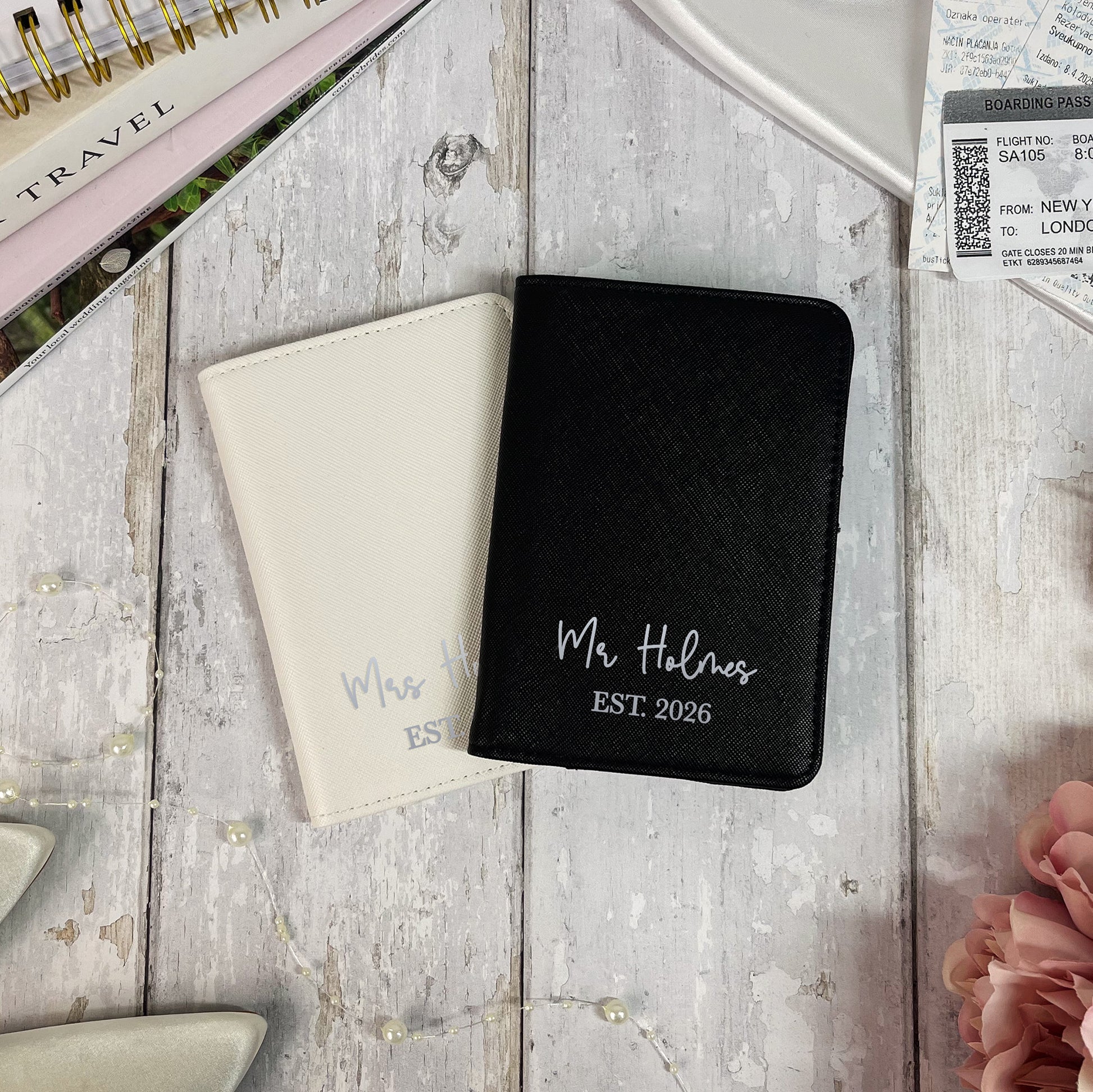 Two personalized passport holders, one black and one beige, on a wooden surface with travel items.