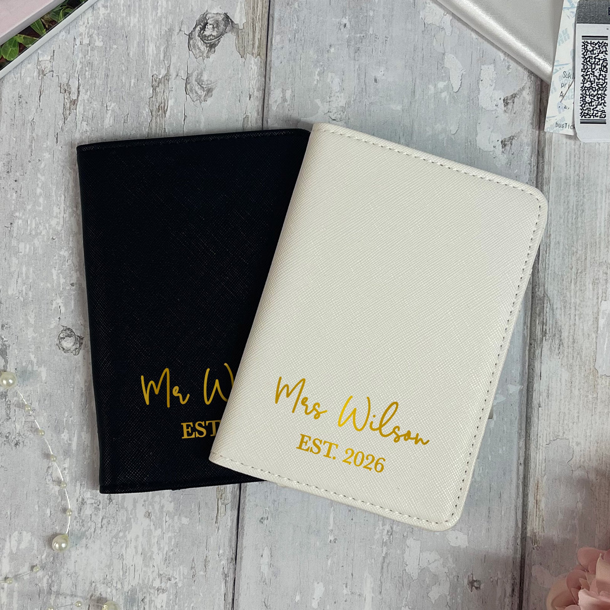 Two personalized passport covers, one black and one white, with 'Mr Wilson' and 'Mrs Wilson EST 2026' text on a wooden surface.