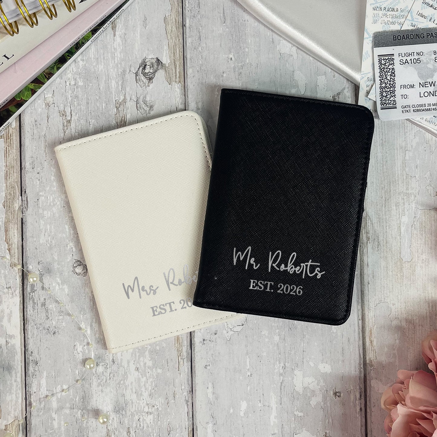 Two personalized passport covers, one black and one beige, on a wooden surface with travel-related items.