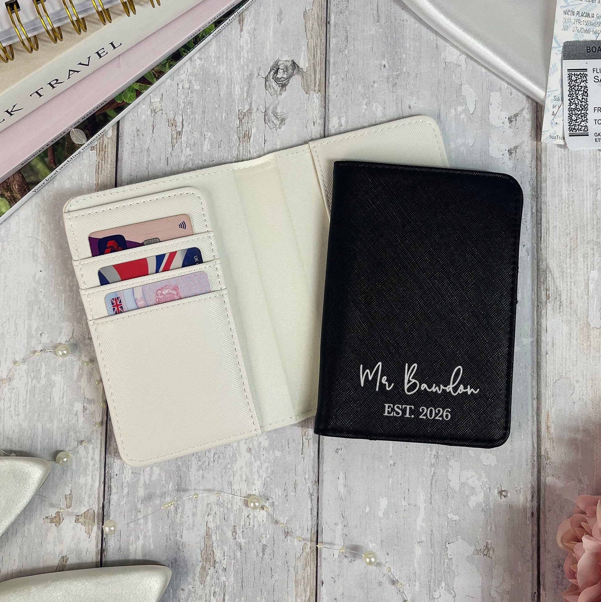 Black passport holder with 'Mr Bawden EST 2026' text on a wooden surface.