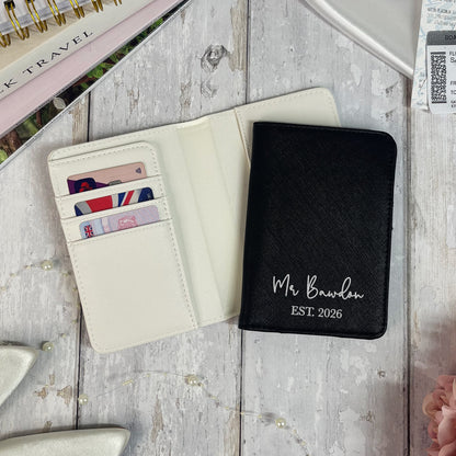 Black passport holder with 'Mr Bawden EST 2026' text on a wooden surface.