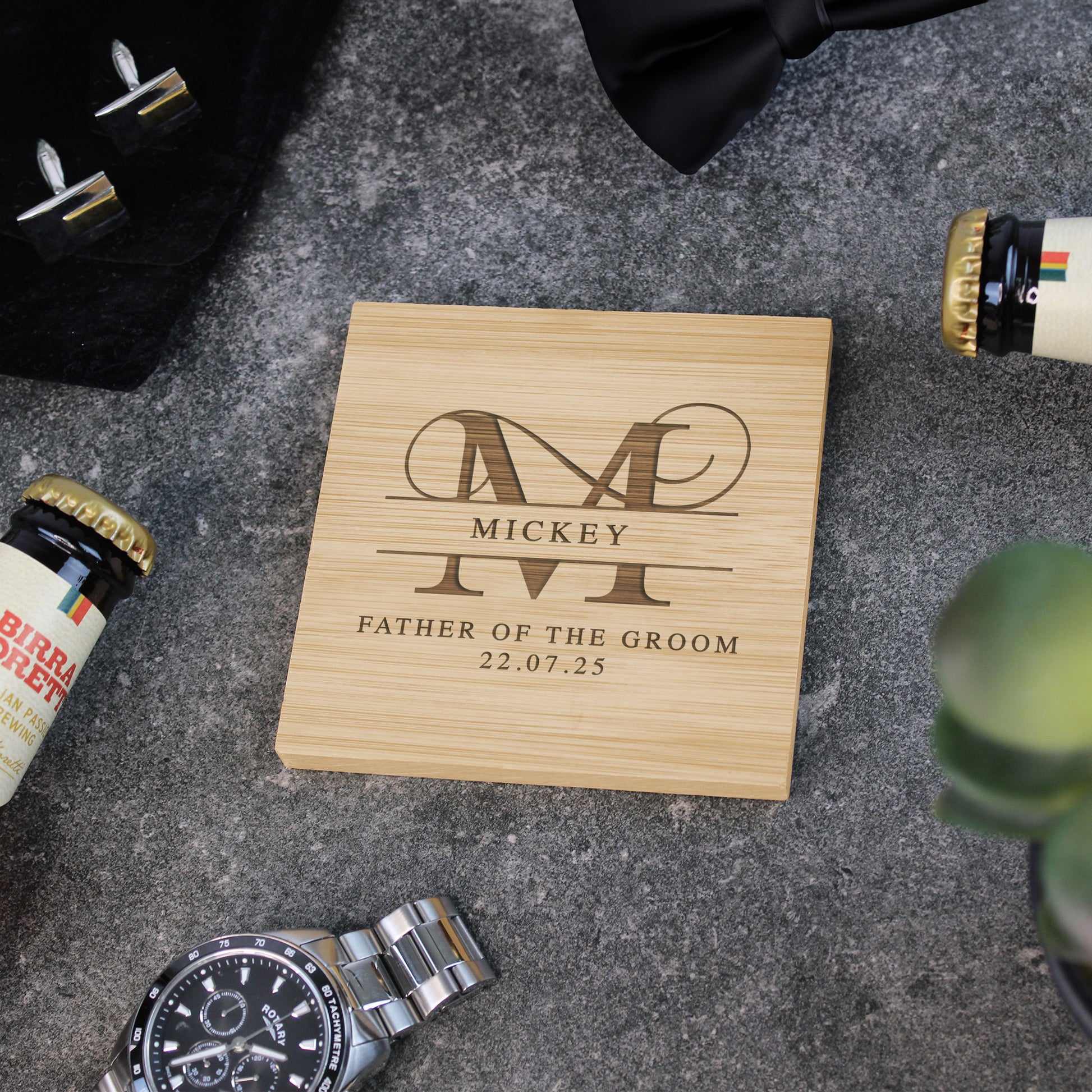 Wooden box with engraved text and monogram on a textured surface with bottles and a watch.