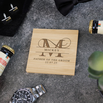 Wooden box with engraved text and monogram on a textured surface with bottles and a watch.