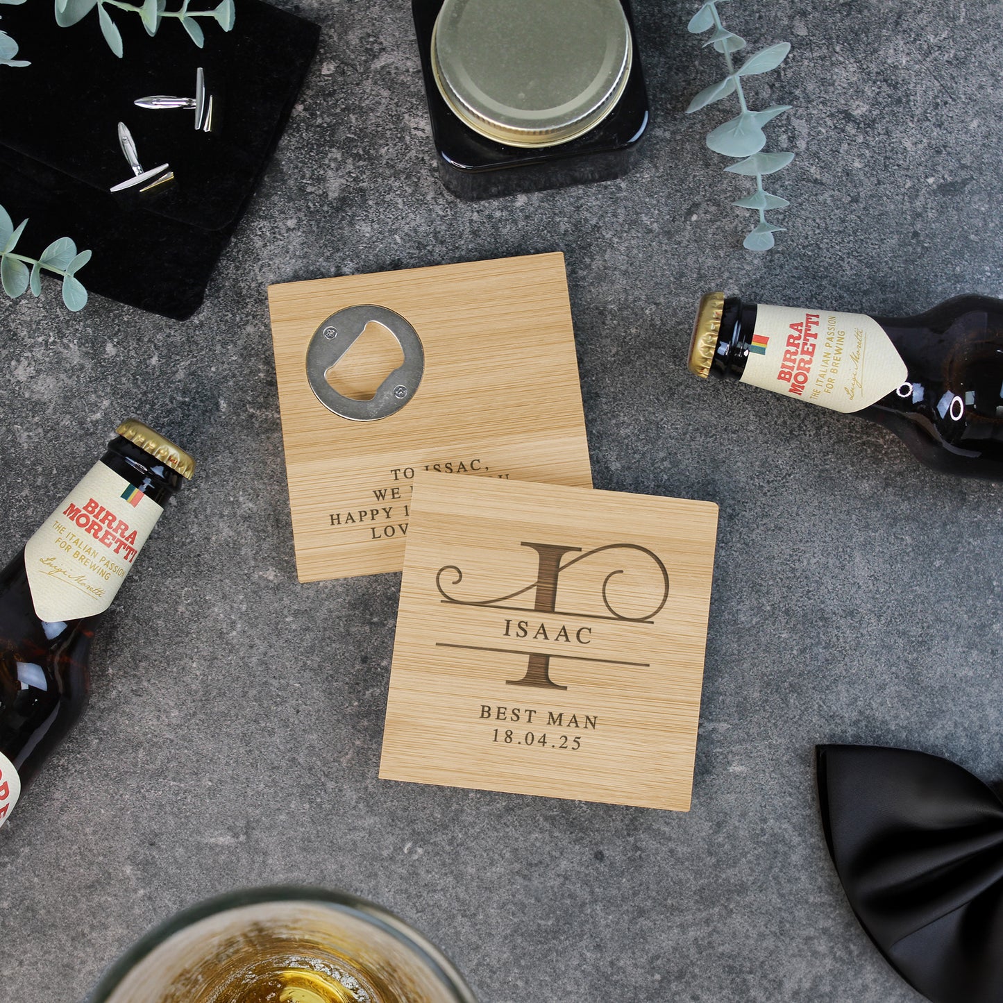 Personalized wooden coasters with engraved names and dates on a gray surface with bottles and a bow tie.
