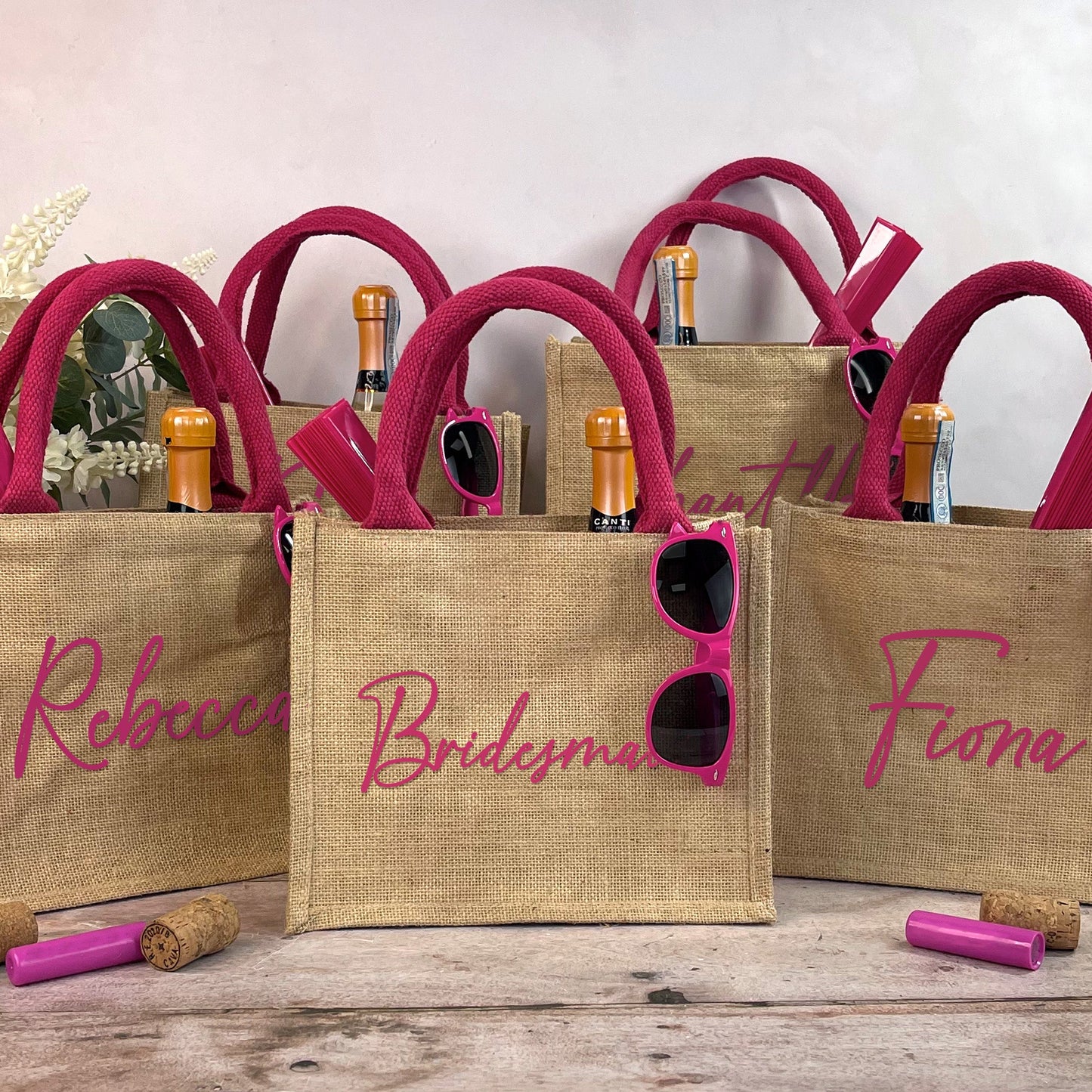 Three jute bags with pink handles, sunglasses, and champagne bottles on a wooden surface.