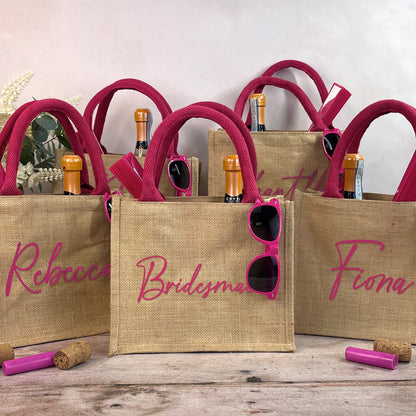 Three jute bags with pink handles, sunglasses, and champagne bottles on a wooden surface.