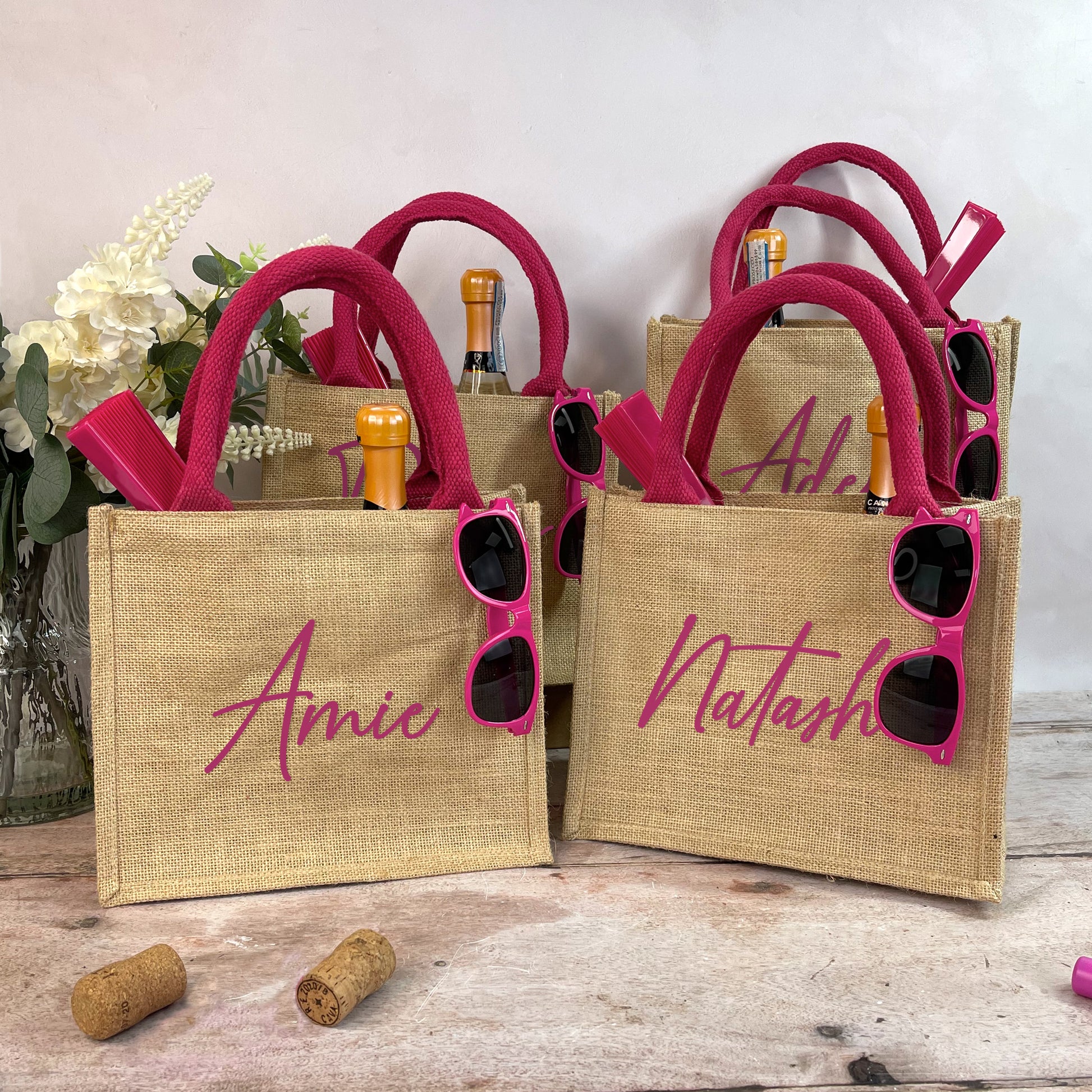 Jute bags with pink handles and personalized names, accompanied by sunglasses and bottles on a wooden surface.