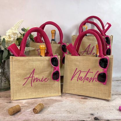 Jute bags with pink handles and personalized names, accompanied by sunglasses and bottles on a wooden surface.