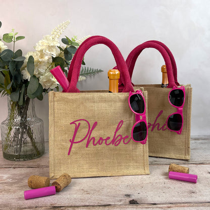 Two jute bags with pink handles and sunglasses, featuring the name 'Phoebe', on a wooden surface with flowers and corks.