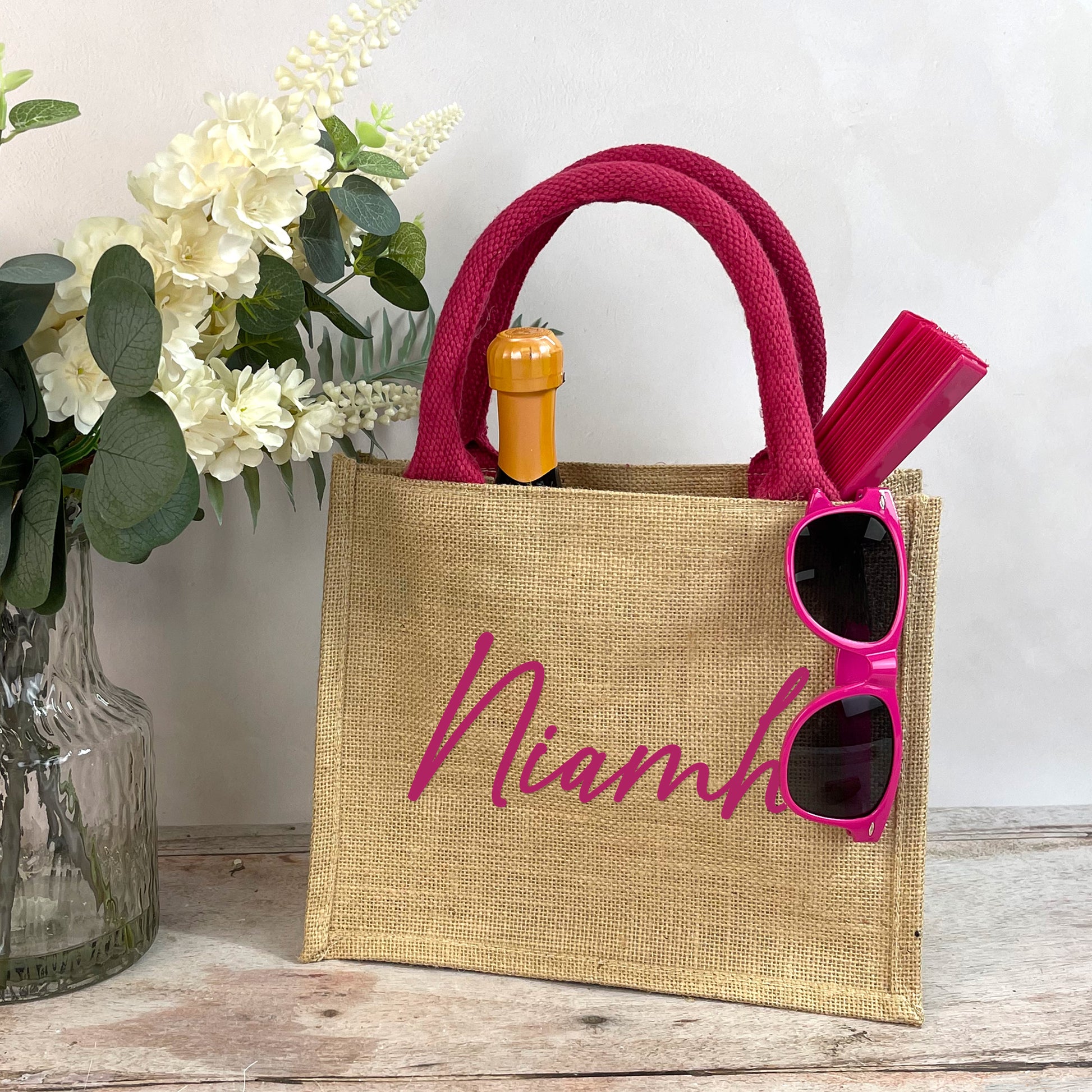 Jute bag with 'Niamh' branding, pink sunglasses, and a bottle on a wooden surface with flowers in the background.